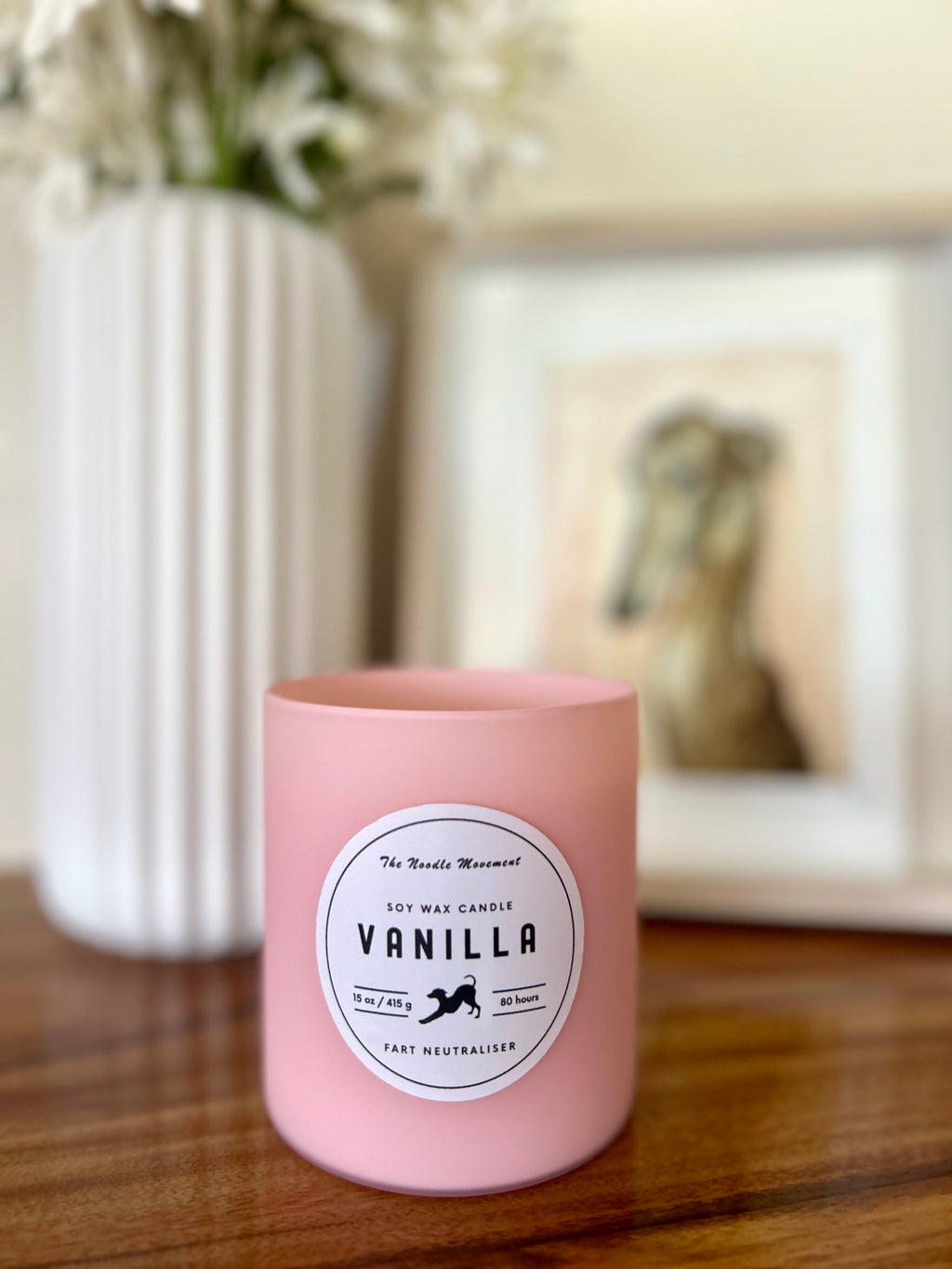 Pink candle labeled 'Vanilla' on a wooden surface with a blurred background